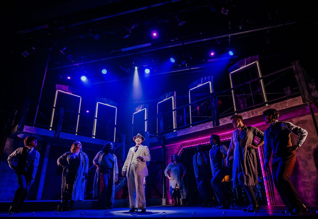 Girl wearing a black and white suit surrounded by 8 more persons, singing during the Chicago play at the Sudbury Theatre