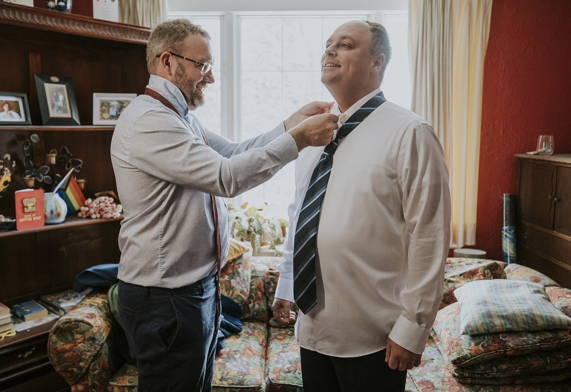 Groom smiling as one of his groomsmen help him with his tie at home in Sudbury