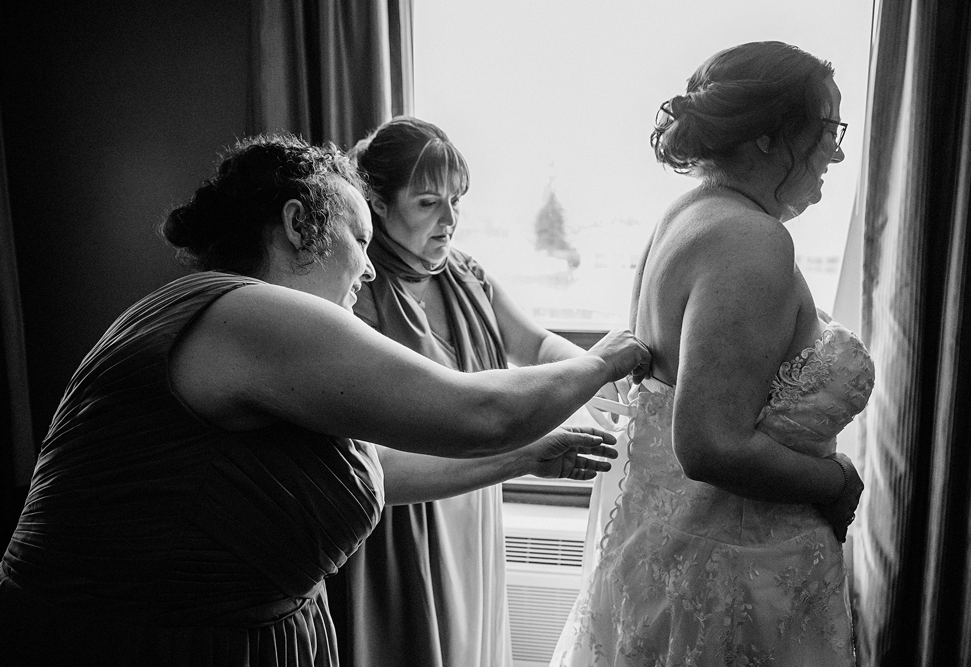 Bride smiling as her bridesmaids help her into her wedding dress at home in Sudbury